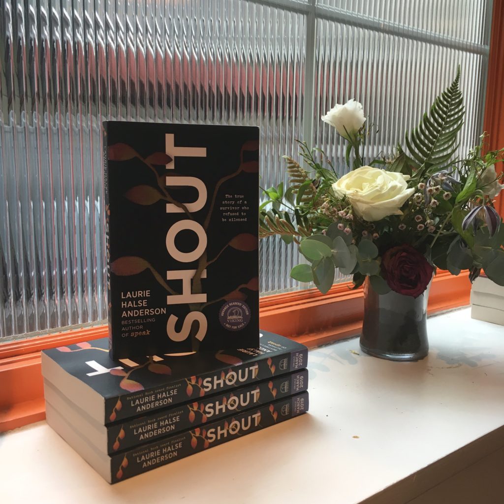 a stack of galleys of SHOUT displayed next to a vase of white roses and ferns